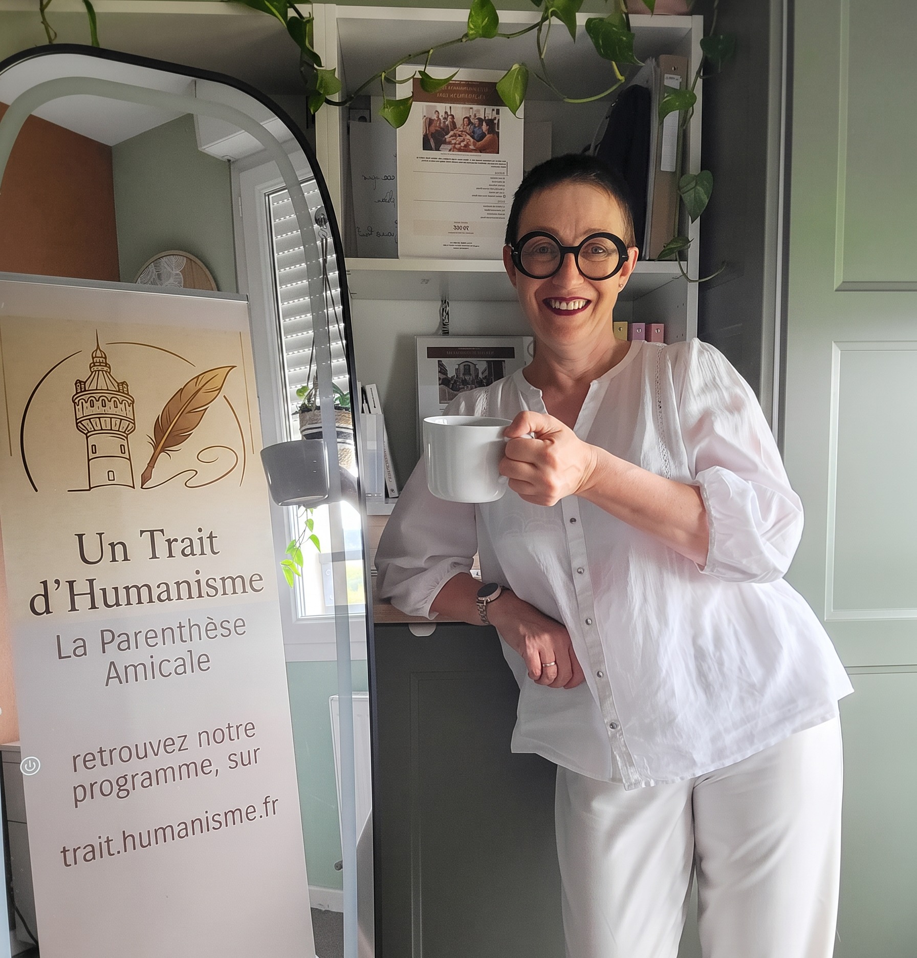 Sylvie Leopold, fondatrice d'Un Trait d'Humanisme, souriante avec une tasse de café devant le kakémono de l'association